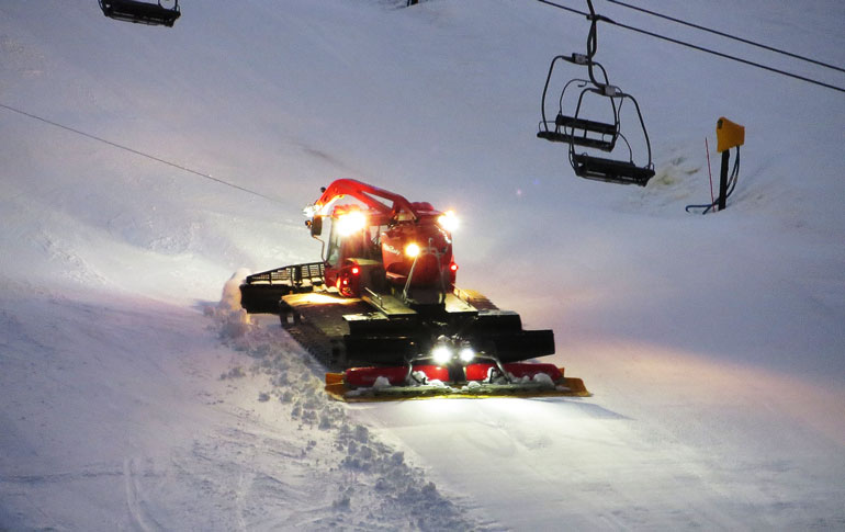 a piste basher grooming the piste at night underneath a chairlift