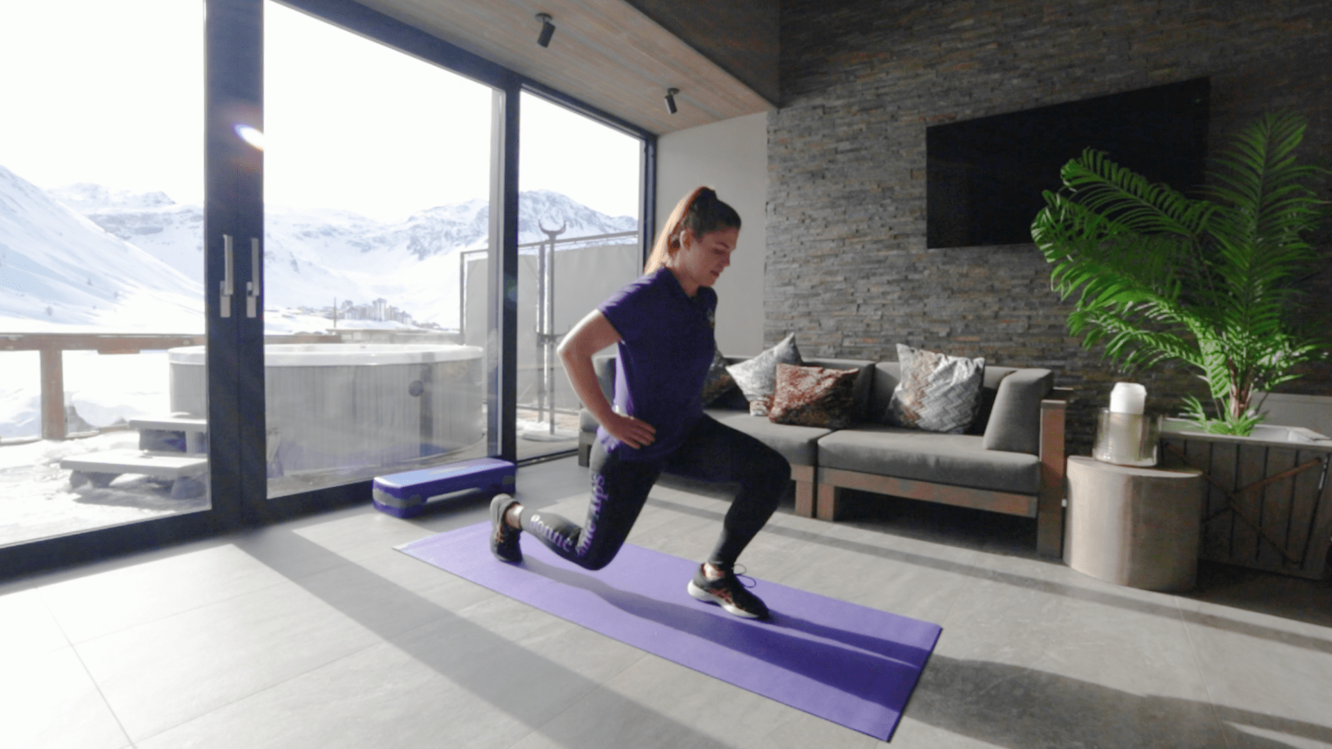 A woman in purple and black gym clothes performs a lunge as part of an at home ski workout
